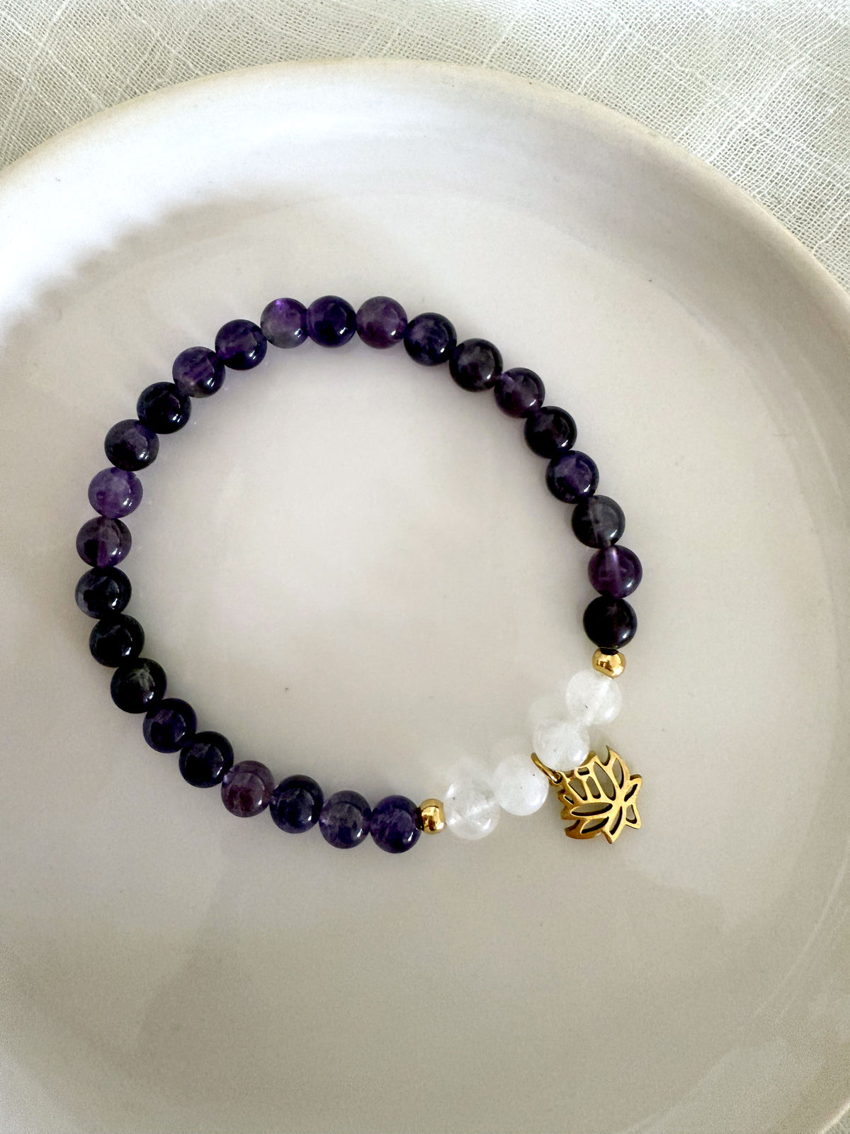 “Pure Consciousness” crown chakra bracelet by Moksha Jewelry, handcrafted with deep amethyst and clear quartz beads, accented with gold-plated details and a lotus charm, displayed on a white ceramic plate—designed to promote spiritual clarity, balance, and awareness.