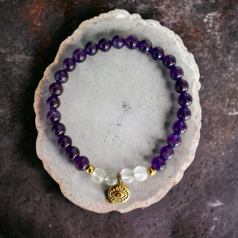 A bracelet made of Amethyst and Rock Crystal Quartz beads, 2 small golden beads and a golden evil eye charm in the middle on a polished gemstone disk.