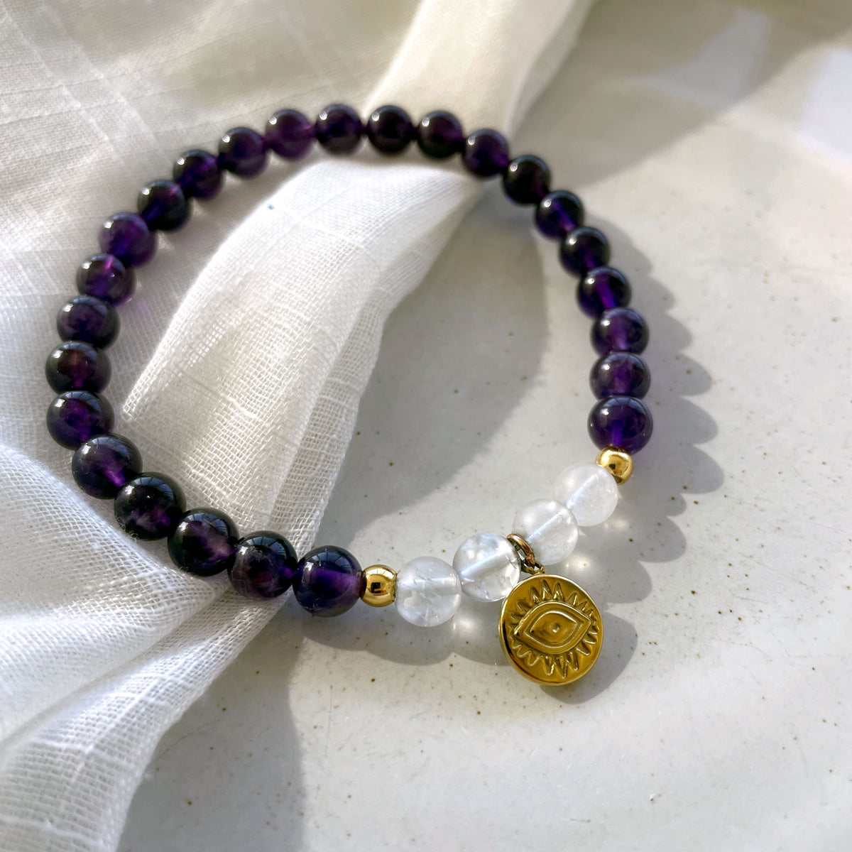 A bracelet made of Amethyst and Rock Crystal Quartz beads, 2 small golden beads and a golden evil eye charm in the middle on a ceramic.