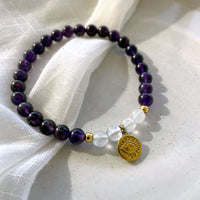 A bracelet made of Amethyst and Rock Crystal Quartz beads, 2 small golden beads and a golden evil eye charm in the middle on a ceramic.