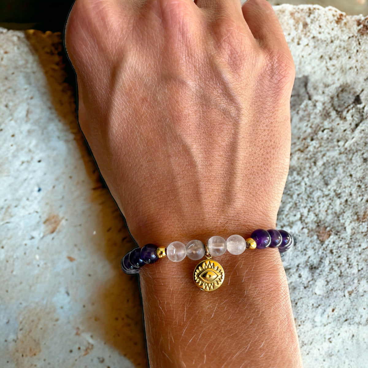 A bracelet made of Amethyst and Rock Crystal Quartz beads, 2 small golden beads and a golden evil eye charm in the middle worn on a wrist.