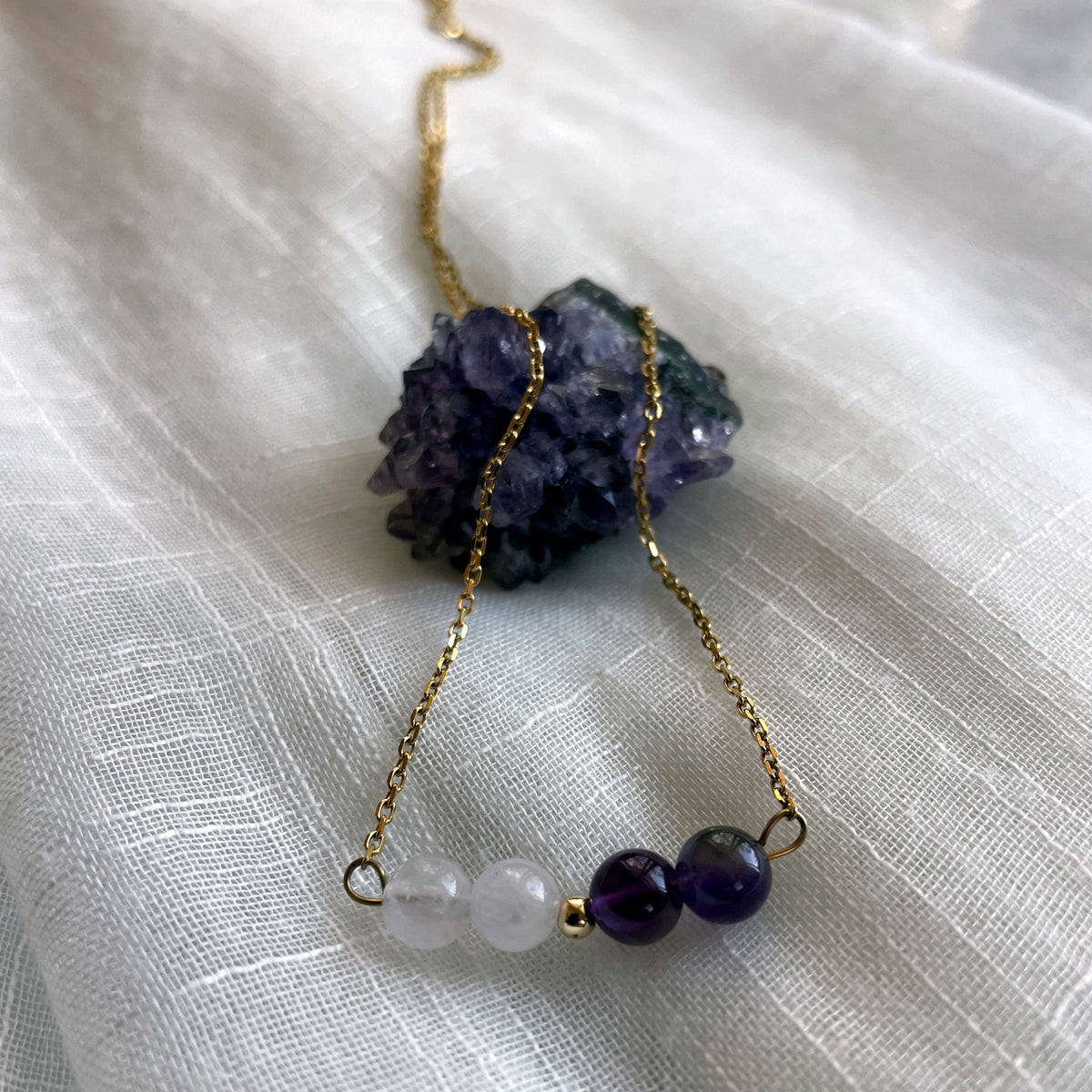 A close-up of a fine golden necklace with 2 Lavender Amethyst beads, 2 Amethyst beads and one golden bead in the middle on a light fabric lying over a raw Amethyst gemstone.