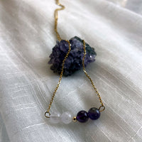 A close-up of a fine golden necklace with 2 Lavender Amethyst beads, 2 Amethyst beads and one golden bead in the middle on a light fabric lying over a raw Amethyst gemstone.