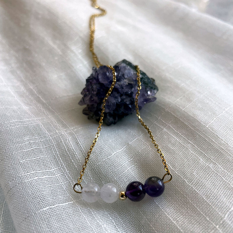 A close-up of a fine golden necklace with 2 Lavender Amethyst beads, 2 Amethyst beads and one golden bead in the middle on a light fabric lying over a raw Amethyst gemstone.