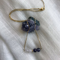A fine golden necklace with 2 Lavender Amethyst beads, 2 Amethyst beads and one golden bead in the middle on a light fabric lying over a raw Amethyst gemstone.