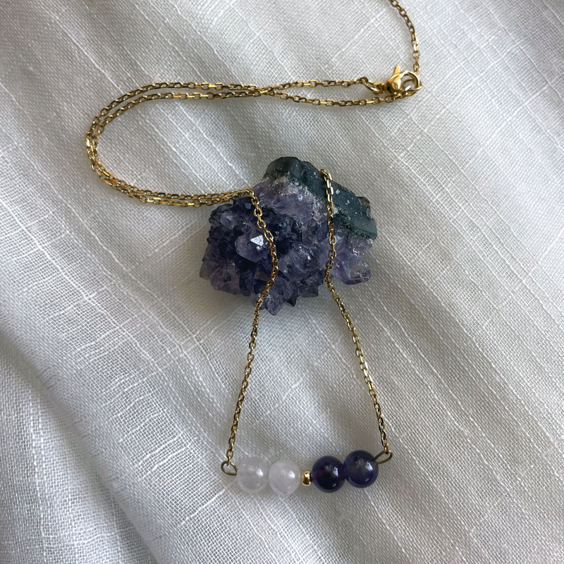 A fine golden necklace with 2 Lavender Amethyst beads, 2 Amethyst beads and one golden bead in the middle on a light fabric lying over a raw Amethyst gemstone.