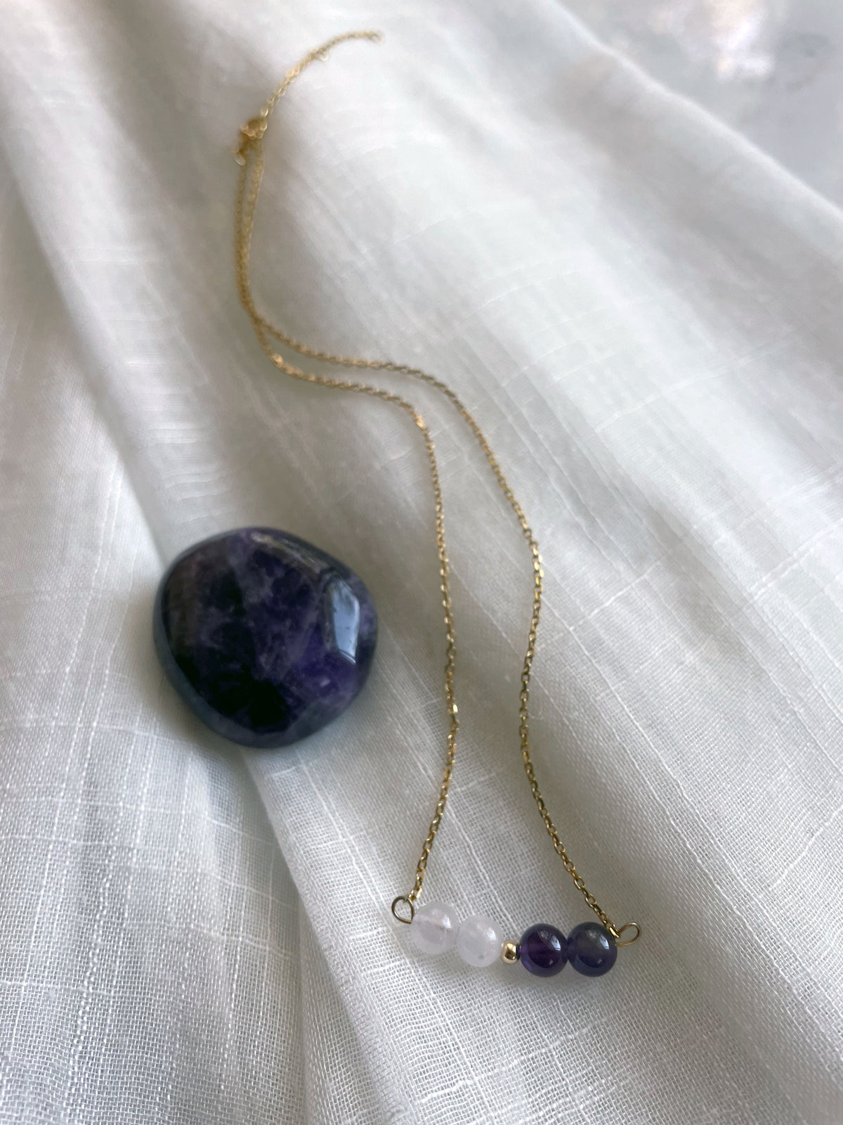 A fine golden necklace with 2 Lavender Amethyst beads, 2 Amethyst beads and one golden bead in the middle on a light fabric next to an Amethyst gemstone.