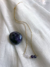 A fine golden necklace with 2 Lavender Amethyst beads, 2 Amethyst beads and one golden bead in the middle on a light fabric next to an Amethyst gemstone.