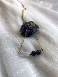 A close-up of a fine golden necklace with 2 Lavender Amethyst beads, 2 Amethyst beads and one golden bead in the middle on a light fabric lying over a raw Amethyst gemstone.