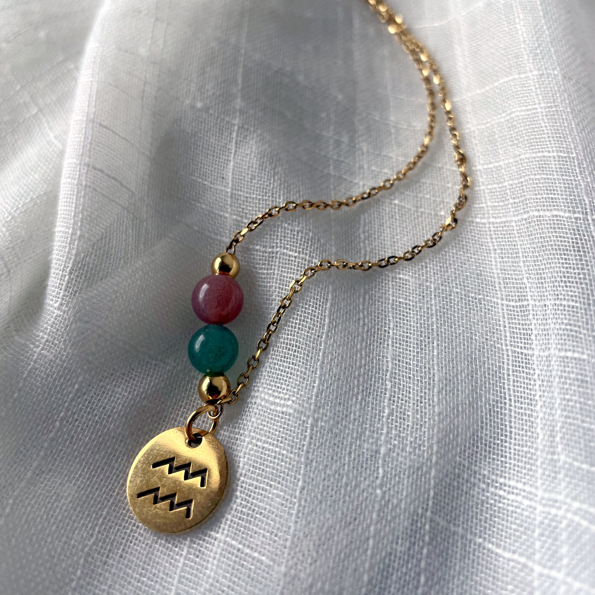 A close-up of a gold plated necklace with a Moonstone bead and a green Aventurine bead, and a gold plated charm with the Aquarius Zodiac sign, on a white linen fabric.