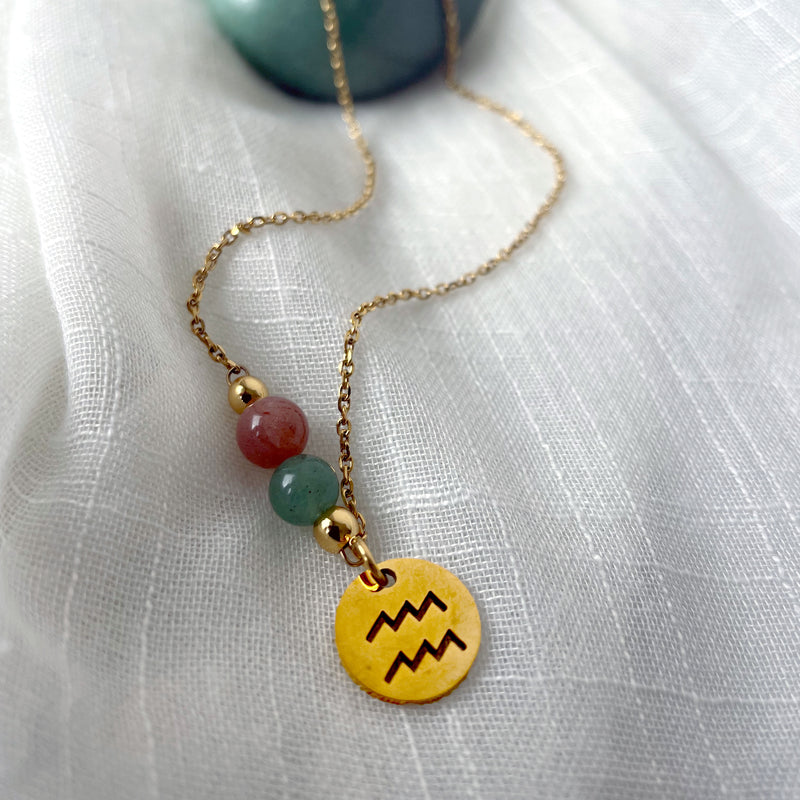 A gold plated necklace with a Moonstone bead and a green Aventurine bead, and a gold plated charm with the Aquarius Zodiac sign, on a white linen fabric.