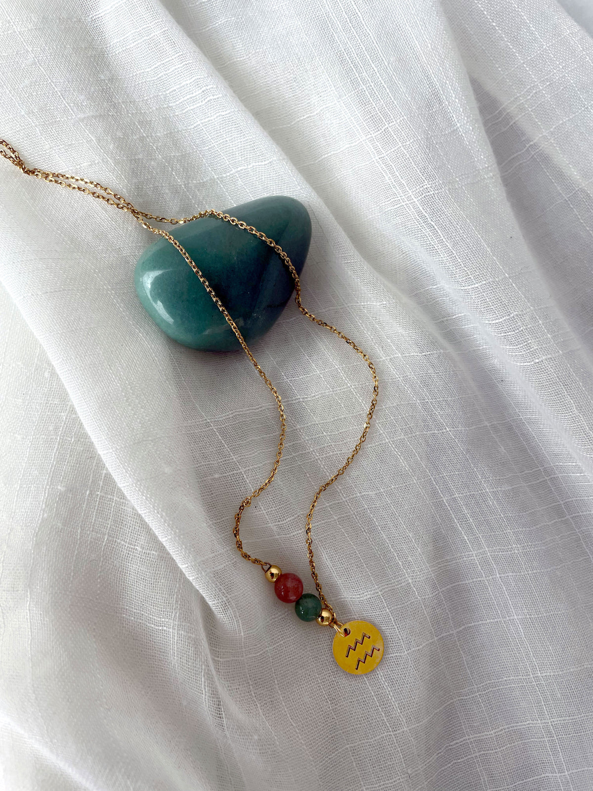 A gold plated necklace with a Moonstone bead and a green Aventurine bead, and a gold plated charm with the Aquarius Zodiac sign, on a white linen fabric.