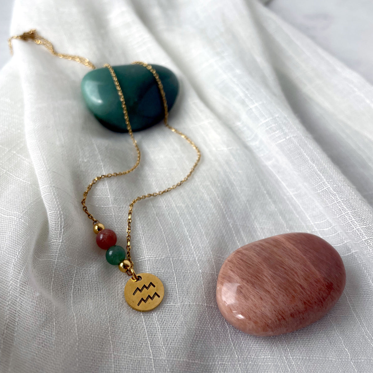 A gold plated necklace with a Moonstone bead and a green Aventurine bead, and a gold plated charm with the Aquarius Zodiac sign, on a white linen fabric.