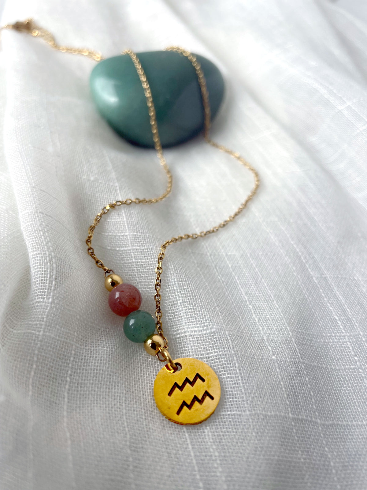 A gold plated necklace with a Moonstone bead and a green Aventurine bead, and a gold plated charm with the Aquarius Zodiac sign, on a white linen fabric.