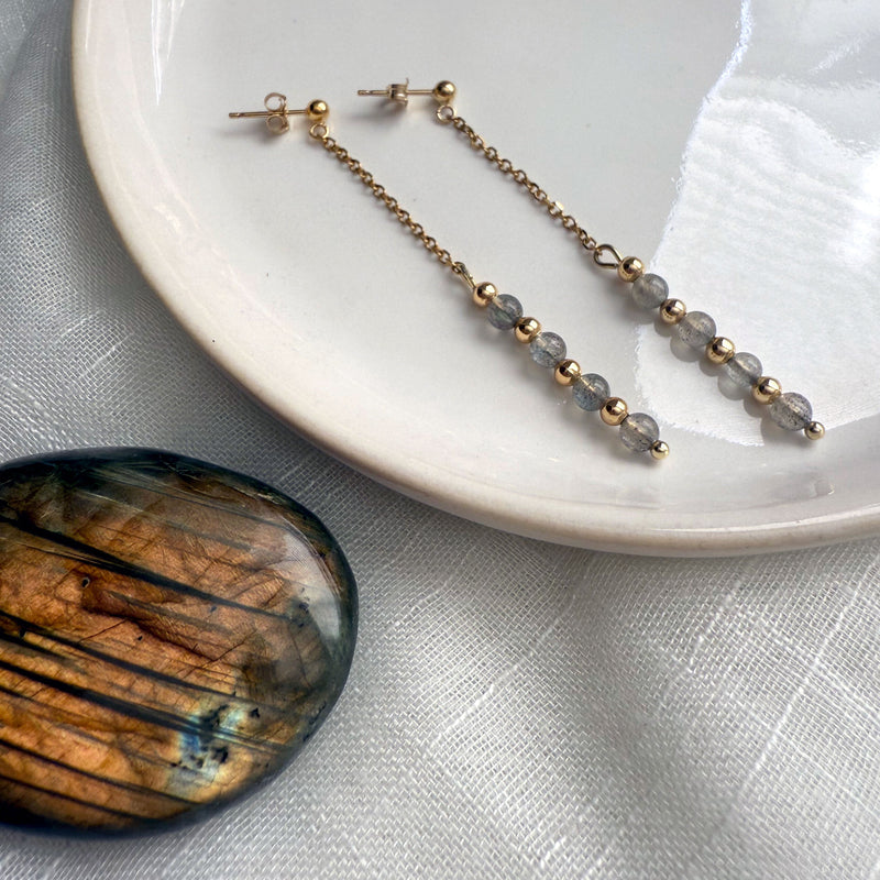 Pair of Moksha Jewelry Aurora Drops on a white ceramic plate beside a labradorite gemstone, featuring gold-filled studs, fine chain, and faceted labradorite beads that reflect light.