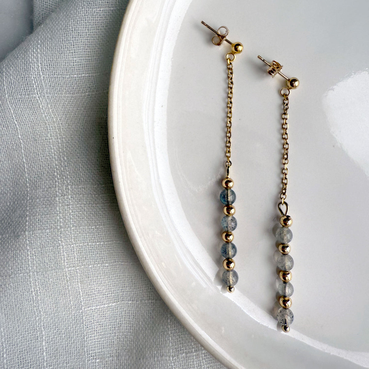 Minimalist product photo of Moksha Jewelry Aurora Drops dangle earrings made with gold-filled posts and labradorite beads, arranged on a white plate with soft natural lighting.