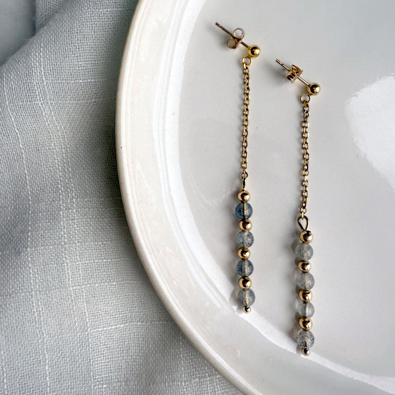 Minimalist product photo of Moksha Jewelry Aurora Drops dangle earrings made with gold-filled posts and labradorite beads, arranged on a white plate with soft natural lighting.