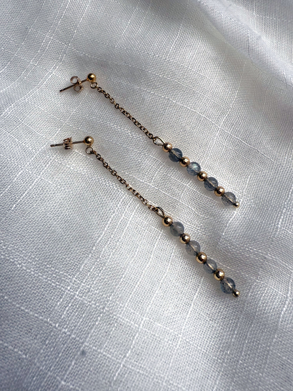 Elegant flat-lay of Moksha Jewelry Aurora Drops earrings on a light linen fabric, showcasing the delicate gold chain, gold-filled findings, and glowing labradorite gemstones.