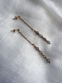 Elegant flat-lay of Moksha Jewelry Aurora Drops earrings on a light linen fabric, showcasing the delicate gold chain, gold-filled findings, and glowing labradorite gemstones.