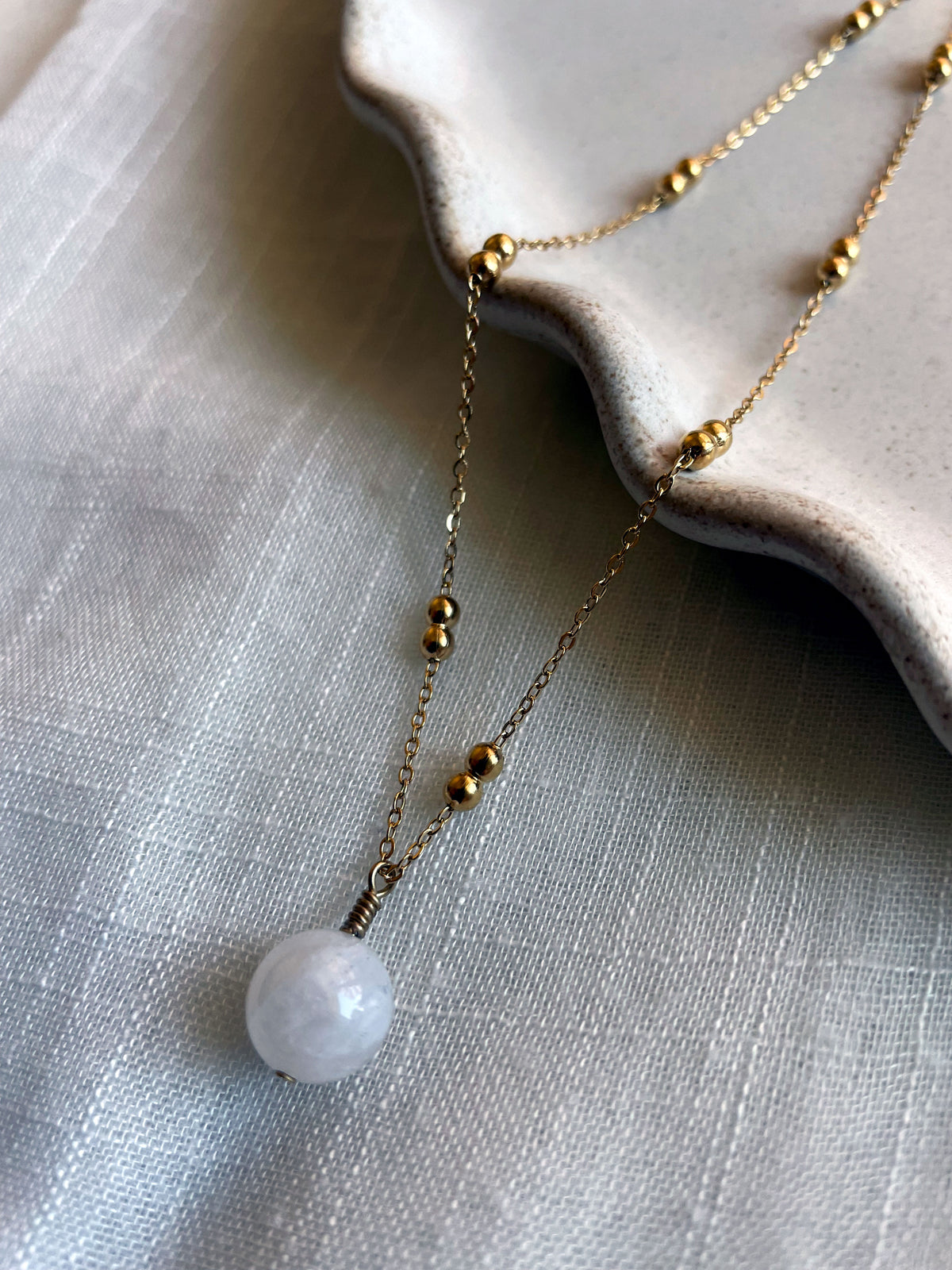 A close-up of a gold plated necklace with a 10mm blue Moonstone bead on a delicate chain with 3mm Ball and 1mm Curb Chain on a ceramic plate on a light fabric.