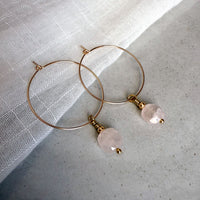 Gold-filled hoop earrings with faceted rose quartz gemstone drops, styled on a soft white linen background.