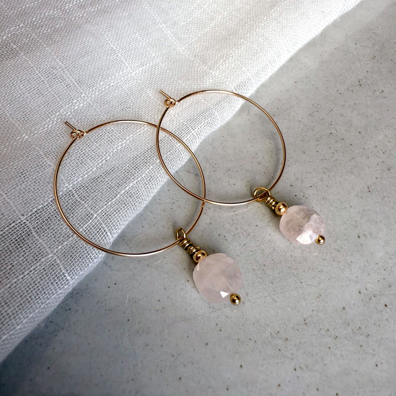 Gold-filled hoop earrings with faceted rose quartz gemstone drops, styled on a soft white linen background.