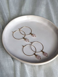 Two pairs of rose quartz hoop earrings with gold accents displayed on a white ceramic dish over soft pastel fabric.