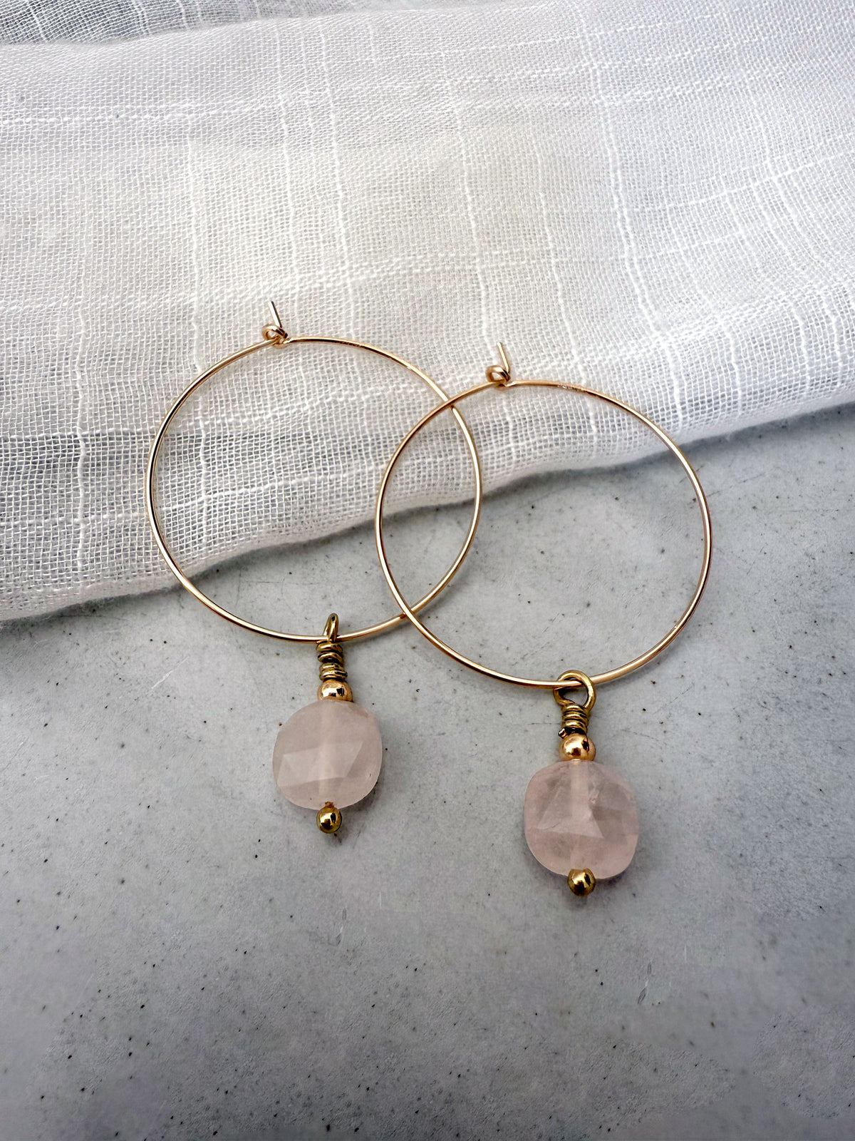 Gold-filled hoop earrings with faceted rose quartz gemstone drops, styled on a soft white linen background.