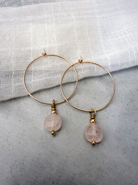 Gold-filled hoop earrings with faceted rose quartz gemstone drops, styled on a soft white linen background.