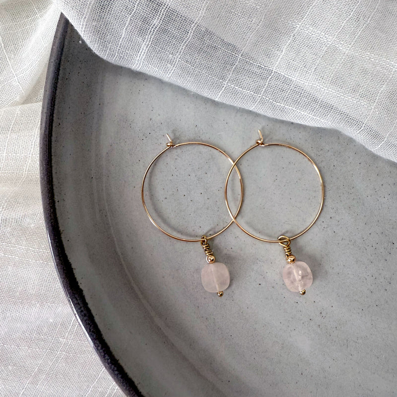 Gold-filled hoop earrings with faceted rose quartz gemstone drops, styled on a soft white linen background.