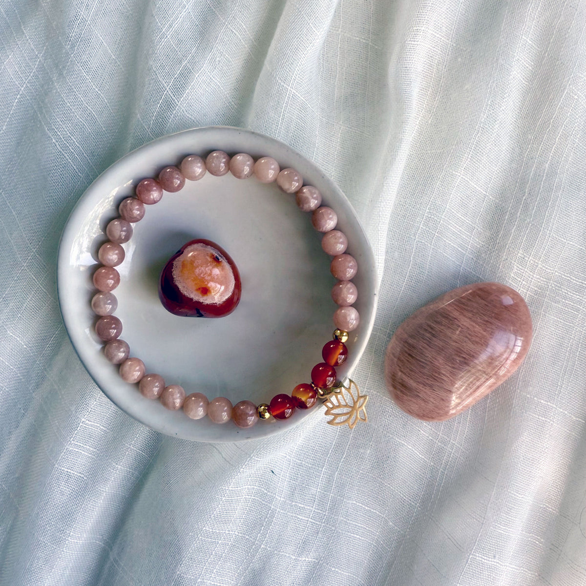 Handcrafted sacral chakra bracelet featuring peach moonstone and carnelian beads with gold accents and a lotus charm, styled on a soft fabric with polished carnelian and moonstone crystals, designed by Moksha Jewelry to inspire passion and joy.