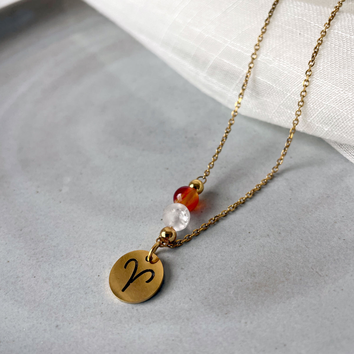 Close-up of a fine necklace with a gold plated Aries Zodiac charm, a Carnelian and a Clear Rock Quartz bead on a fine gold plated chain, laying on a grey ceramic plate.