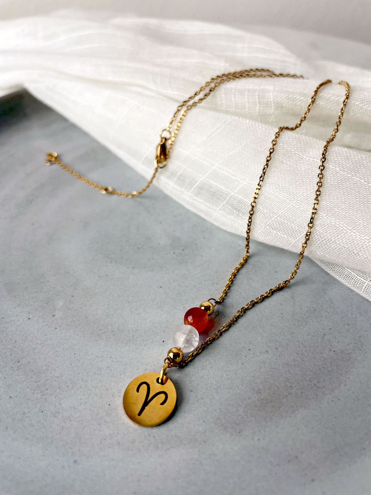 Close-up of a fine necklace with a gold plated Aries Zodiac charm, a Carnelian and a Clear Rock Quartz bead on a fine gold plated chain, laying on a grey ceramic plate.