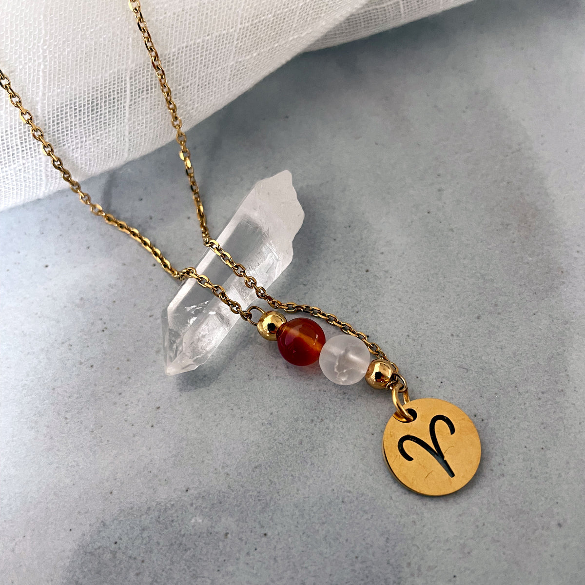 Close-up of a fine necklace with a gold plated Aries Zodiac charm, a Carnelian and a Clear Rock Quartz bead on a fine gold plated chain, laying on a grey ceramic plate.