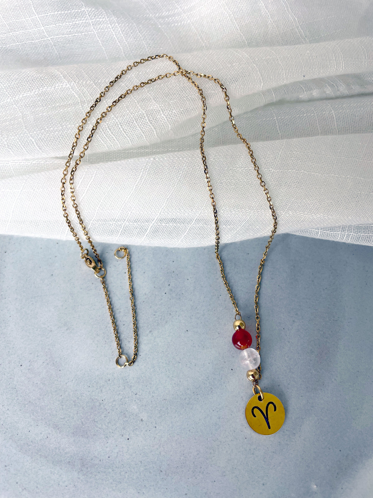 A fine necklace with a gold plated Aries Zodiac charm, a Carnelian and a Clear Rock Quartz bead on a fine gold plated chain, laying on a grey ceramic plate.