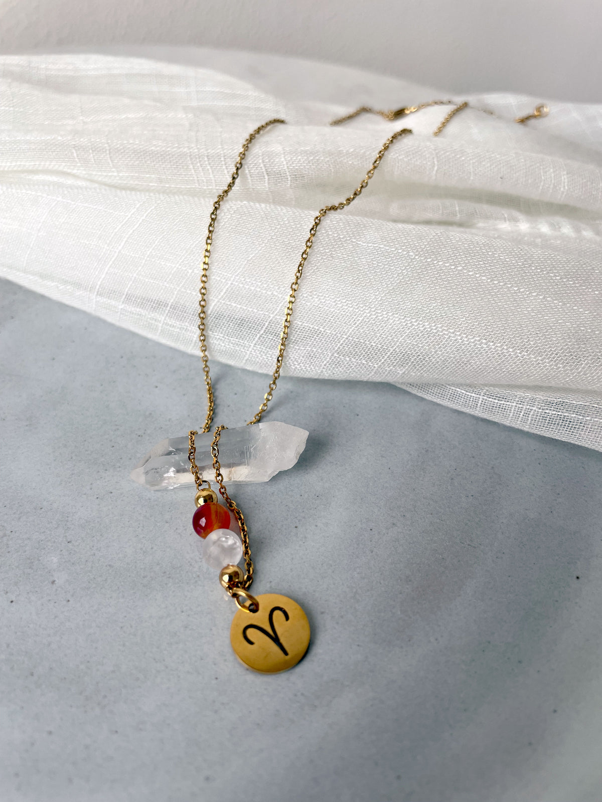 Close-up of a fine necklace with a gold plated Aries Zodiac charm, a Carnelian and a Clear Rock Quartz bead on a fine gold plated chain, laying on a clear quartz tip on a grey ceramic plate.