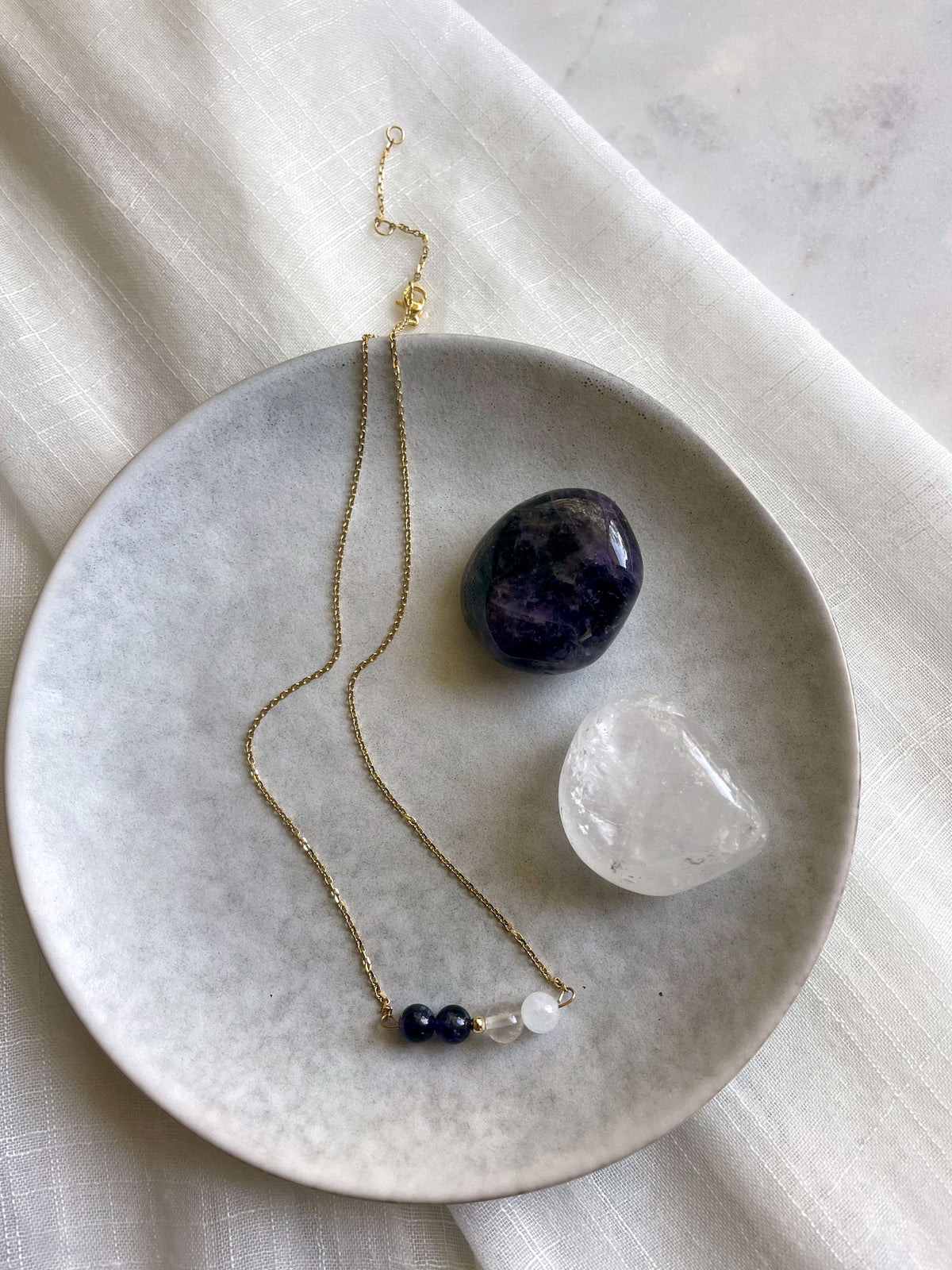 A fine golden necklace with 2 Rock Crystal Quartz beads, one small golden bead and 2 Amethyst beads on a ceramic plate next to a Clear Quartz and Amethyst gemstone.