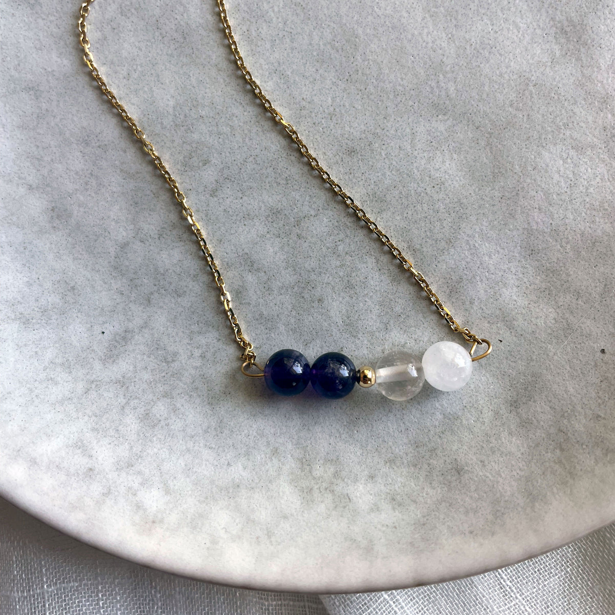 A close-up of a fine golden necklace with 2 Rock Crystal Quartz beads, one small golden bead and 2 Amethyst beads on a ceramic plate.