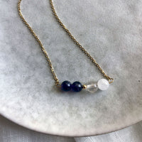 A close-up of a fine golden necklace with 2 Rock Crystal Quartz beads, one small golden bead and 2 Amethyst beads on a ceramic plate.