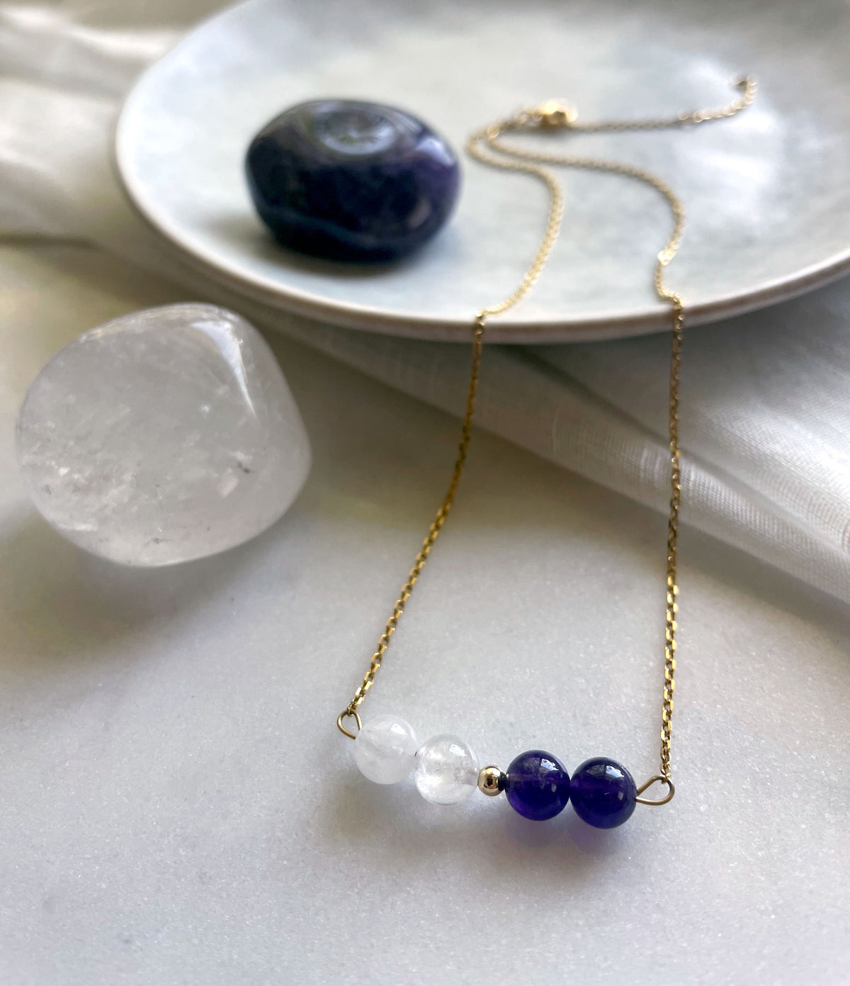 A fine golden necklace with 2 Rock Crystal Quartz beads, one small golden bead and 2 Amethyst beads on a ceramic plate next to a Clear Quartz and Amethyst gemstone.