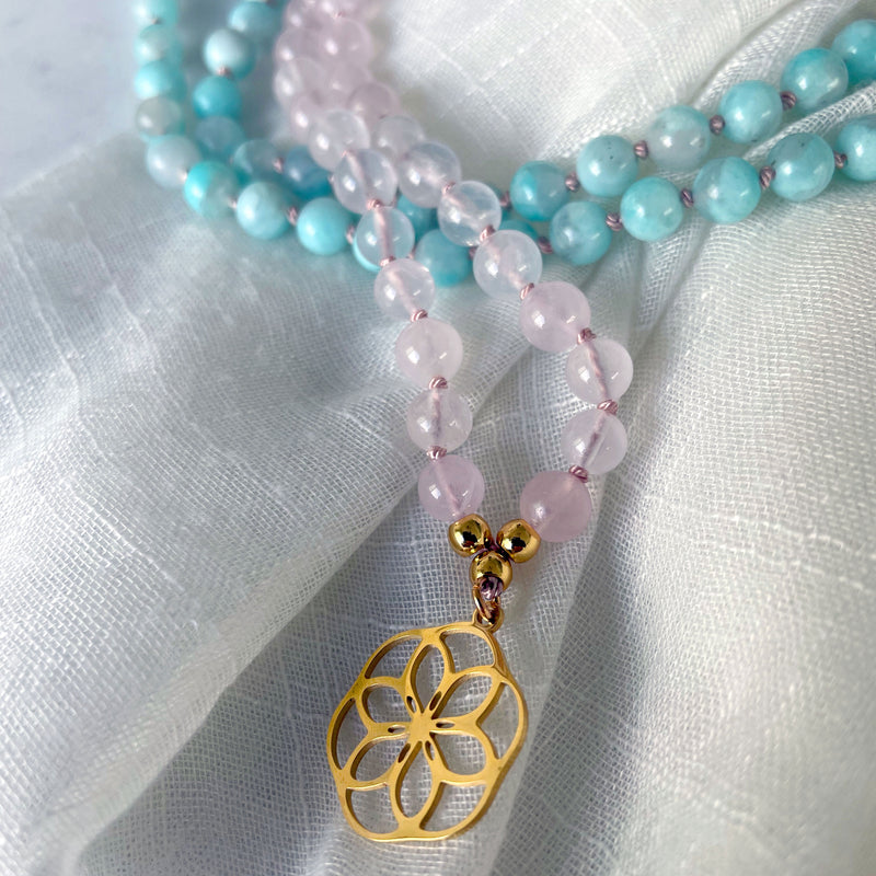 A close-up of a Mala made of 6mm Rose Quartz beads and 6mm Amazonite beads with a gold plated flower pendant on a light fabric.