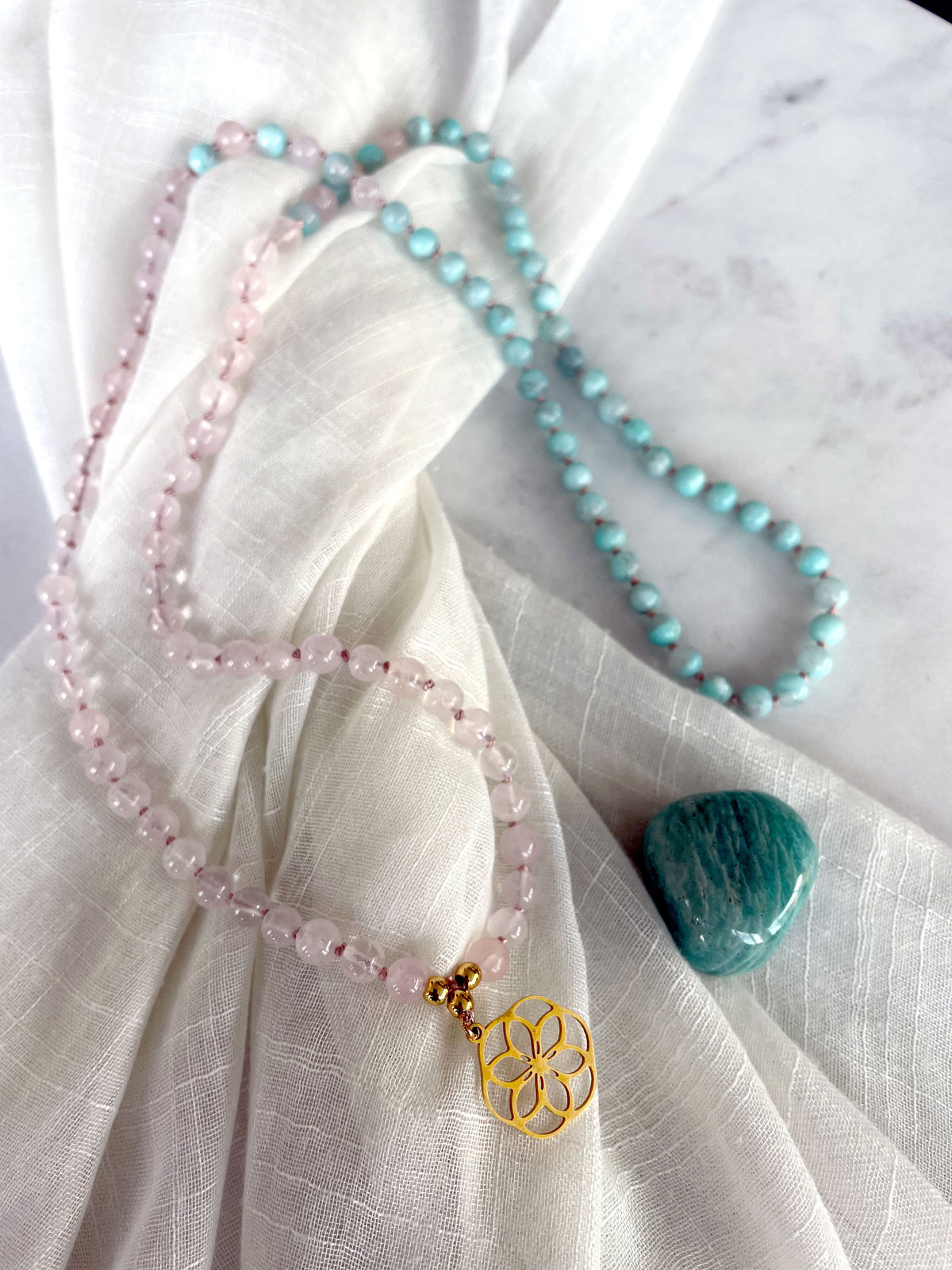 A Mala made of 6mm Rose Quartz beads and 6mm Amazonite beads with a gold plated flower pendant next to a polished Amazonite gemstone on a light fabric.