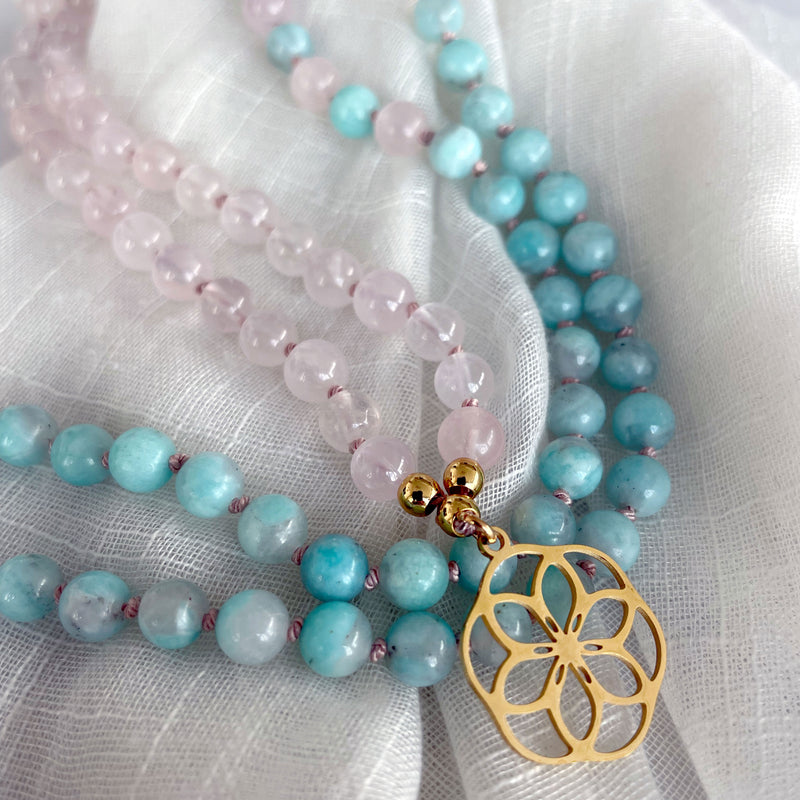 A close-up of a Mala made of 6mm Rose Quartz beads and 6mm Amazonite beads with a gold plated flower pendant on a light fabric.