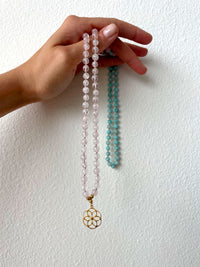 A hand holds a Mala made of 6mm Rose Quartz beads and 6mm Amazonite beads with a gold plated flower pendant.