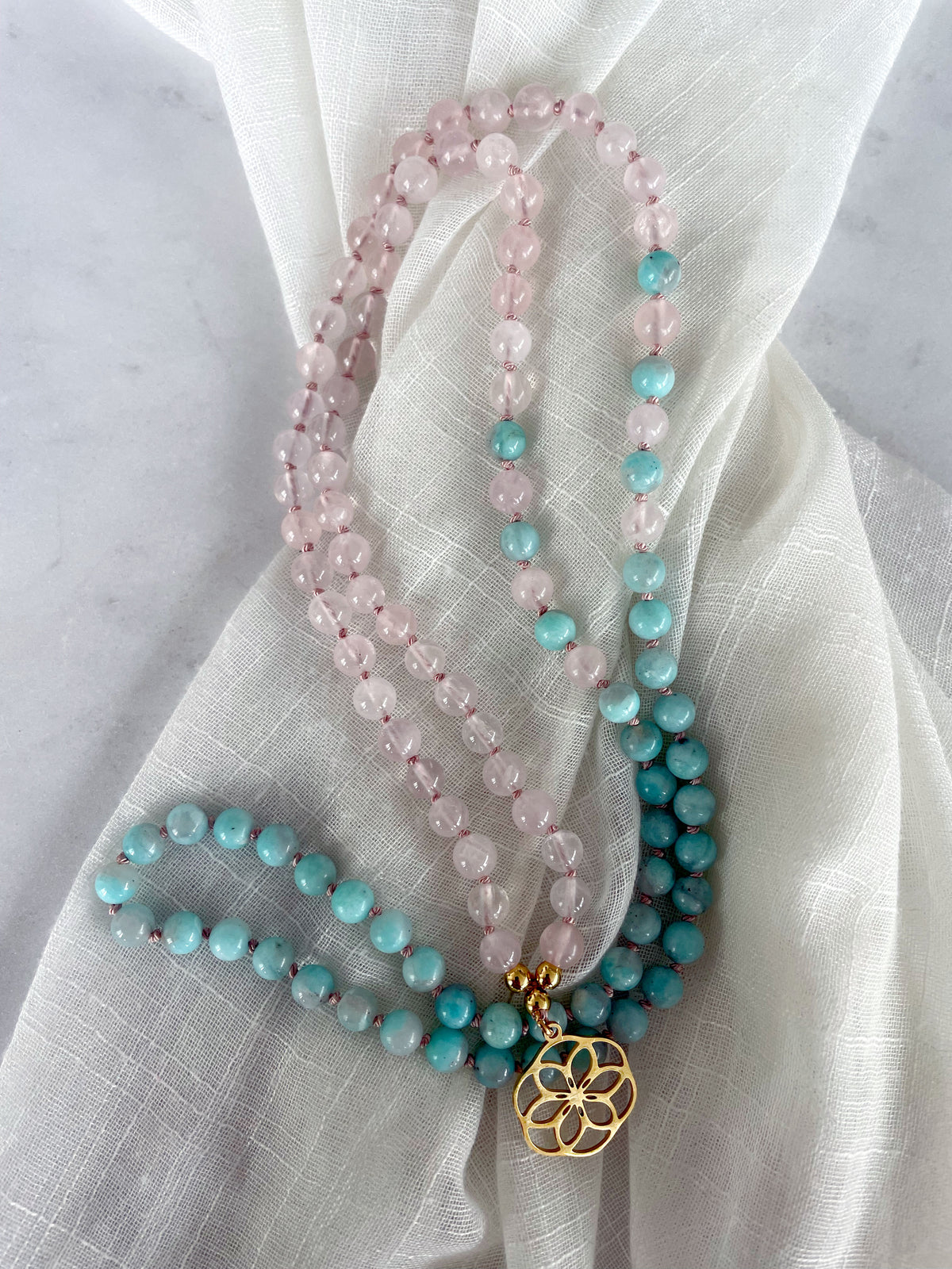 A Mala made of 6mm Rose Quartz beads and 6mm Amazonite beads with a gold plated flower pendant on a light fabric.