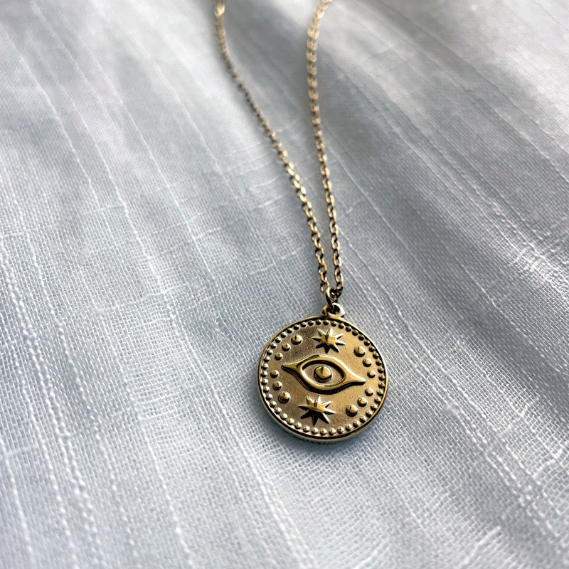 A close-up of a fine gold plated stainless steel necklace with an Evil Eye pendant on a light fabric.