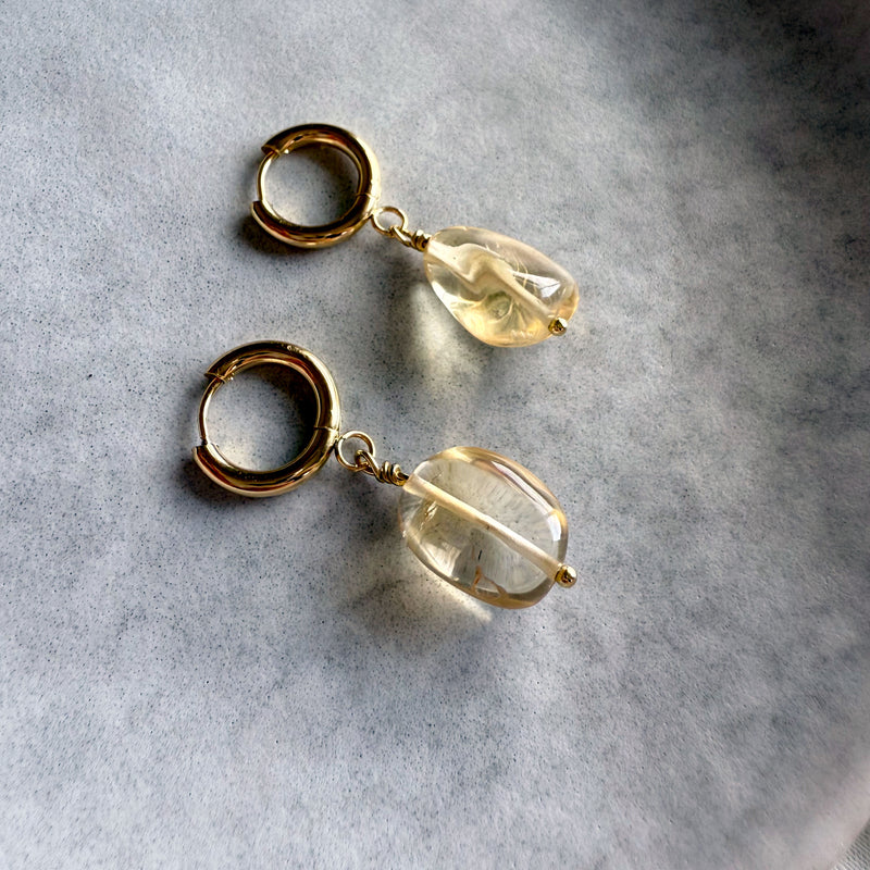 Gold-plated huggie hoop earrings with citrine gemstone drops, displayed on a soft gray background. The citrine stones are translucent with natural inclusions, reflecting warm golden hues.