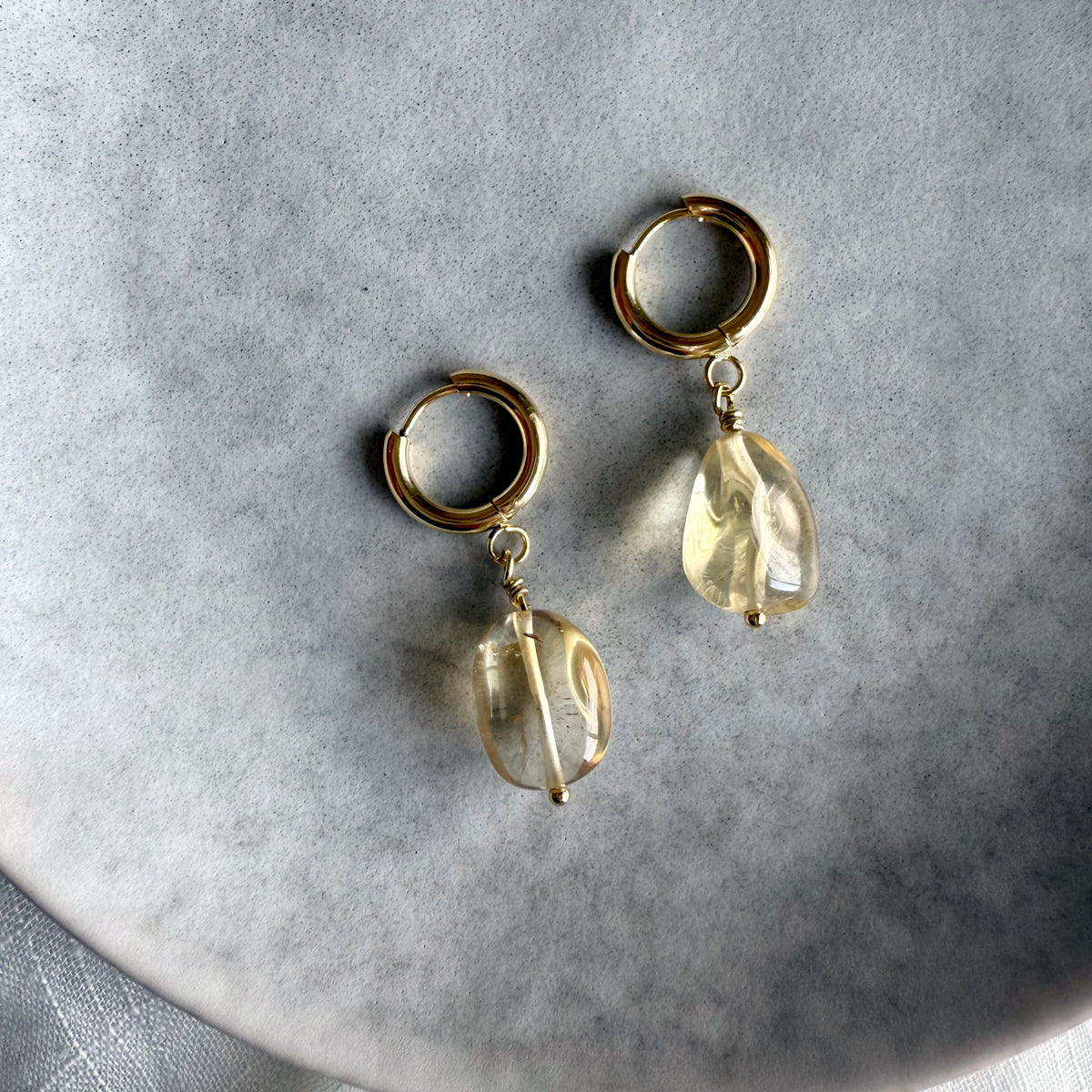 Gold-plated huggie hoop earrings with citrine gemstone drops, displayed on a soft gray background. The citrine stones are translucent with natural inclusions, reflecting warm golden hues.