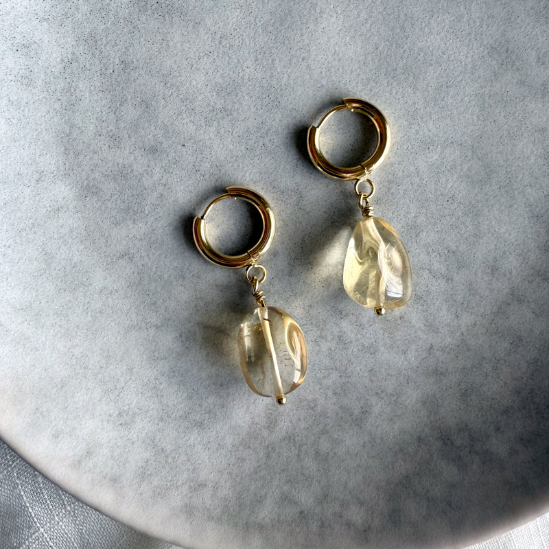 Gold-plated huggie hoop earrings with citrine gemstone drops, displayed on a soft gray background. The citrine stones are translucent with natural inclusions, reflecting warm golden hues.
