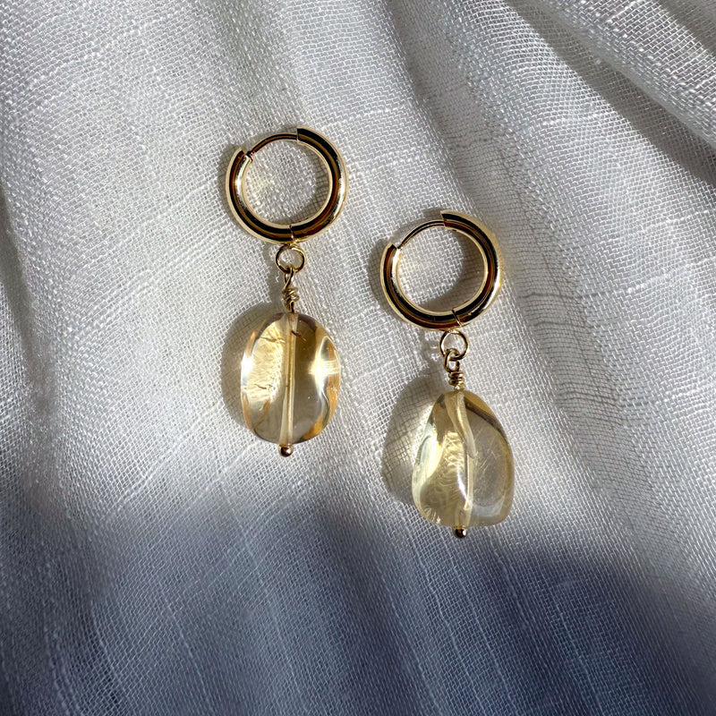 Gold-plated huggie hoop earrings with citrine gemstone drops, elegantly displayed on softly draped white fabric. Sunlight highlights the gemstones’ natural inclusions and golden glow, casting delicate shadows.